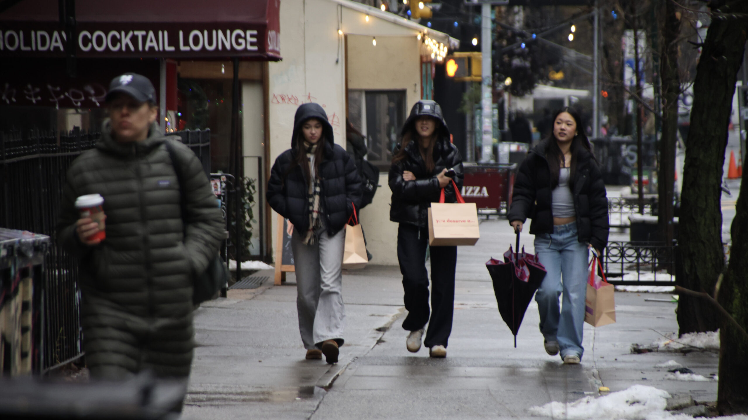 This is a candid 16:9 street photo taken on a wet, overcast winter day, probably in a dense city neighborhood