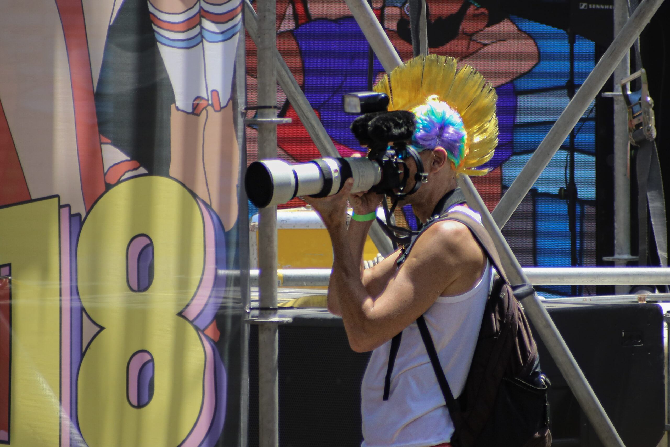 Photographer in a gold mohawk wig and white tank top shooting with a white telephoto lens in front of a pride parade stage with cartoon banners and scaffolding