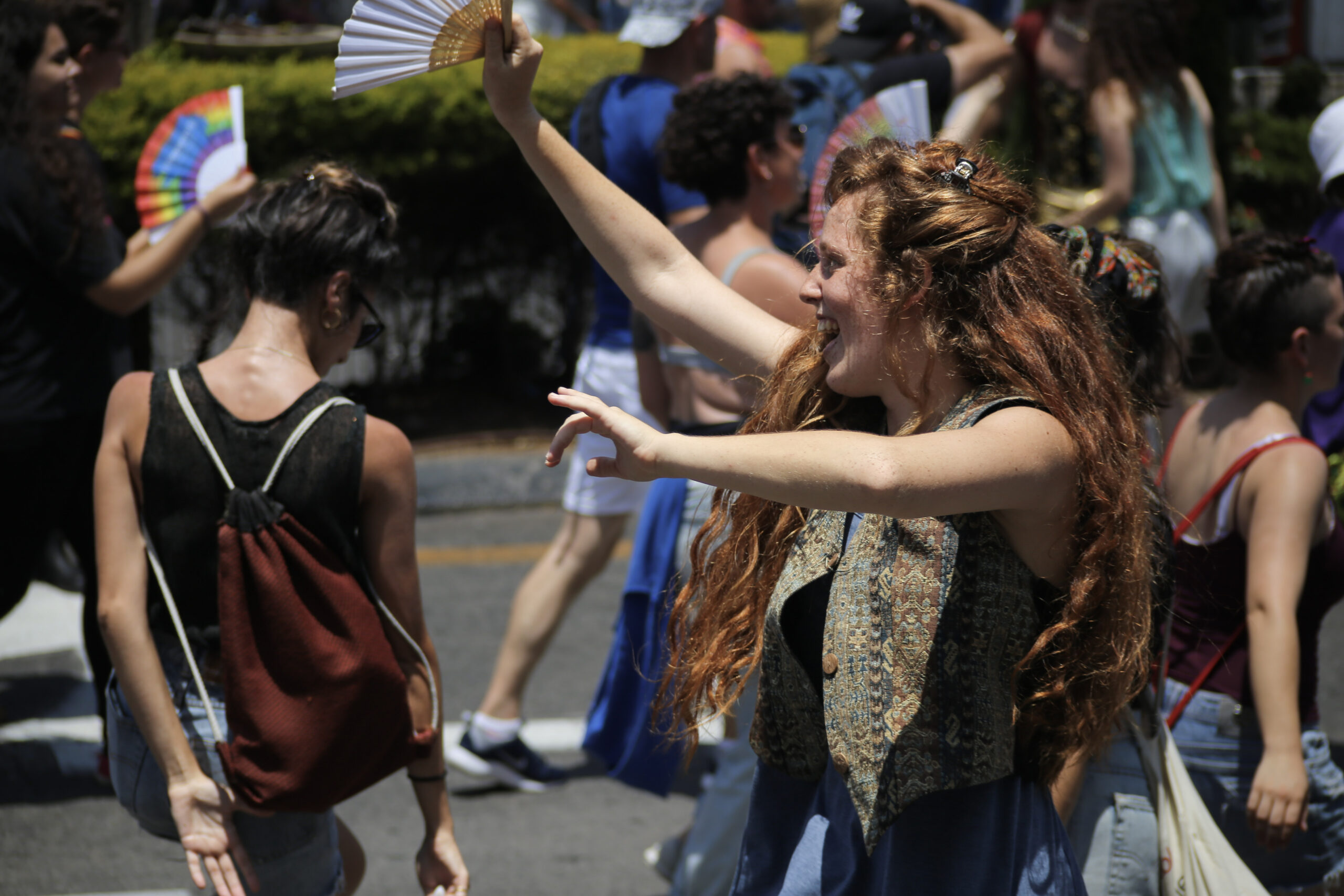 Woman with long red curly hair and a tapestry vest laughing with a white fan raised overhead in a crowded pride parade, rainbow fans in the background