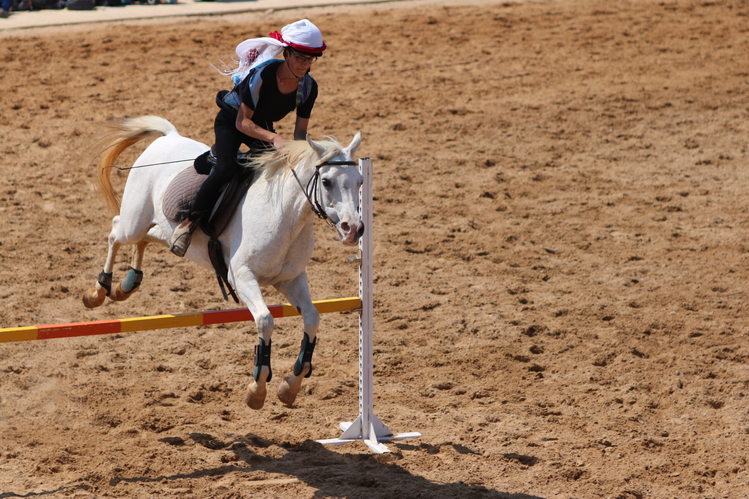 Grey horse and rider in a white headscarf jumping an orange and yellow rail in a sand arena