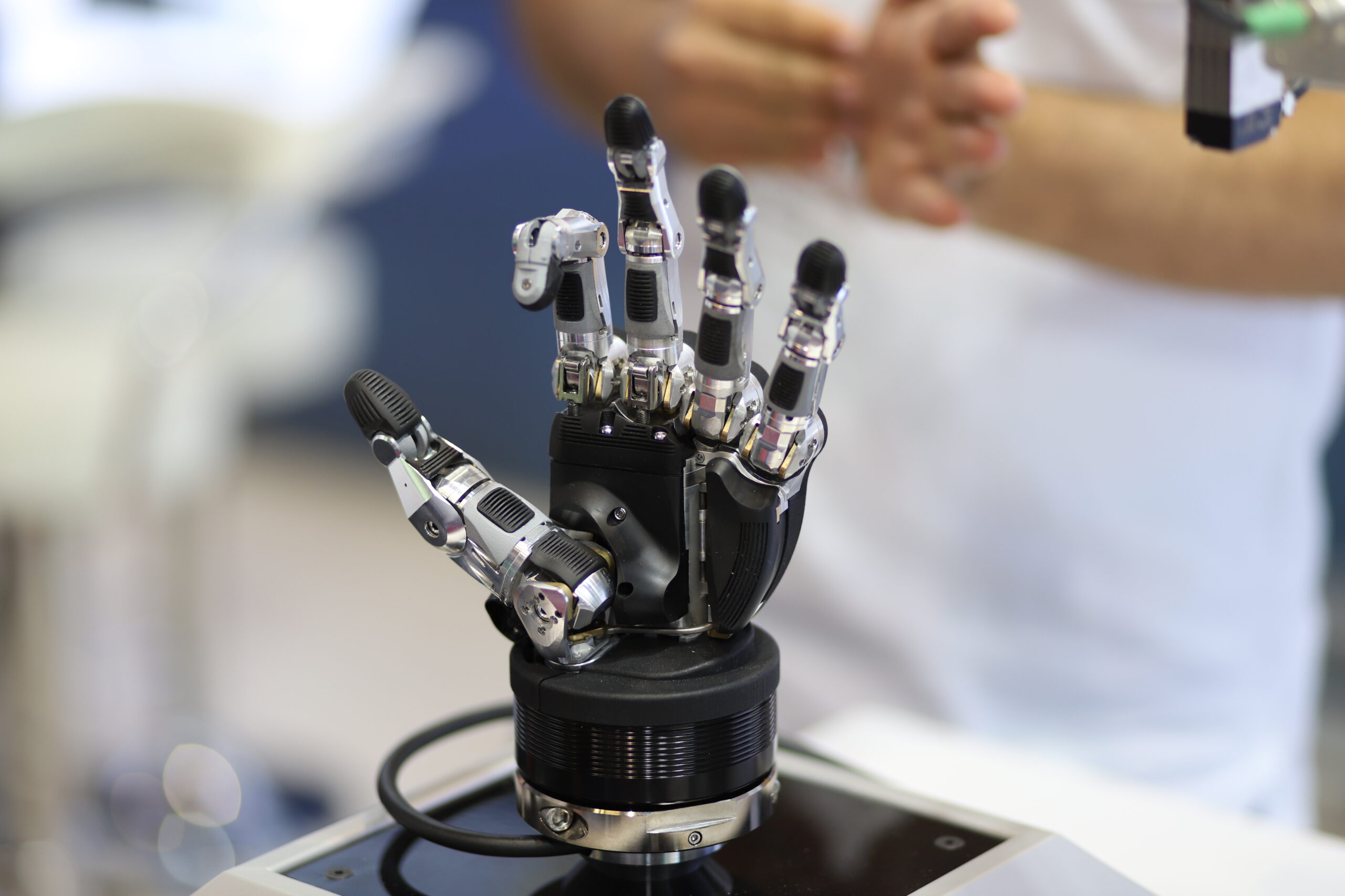 Dexterous robotic hand with five articulated metal fingers mounted on a black actuator base at a trade show stand