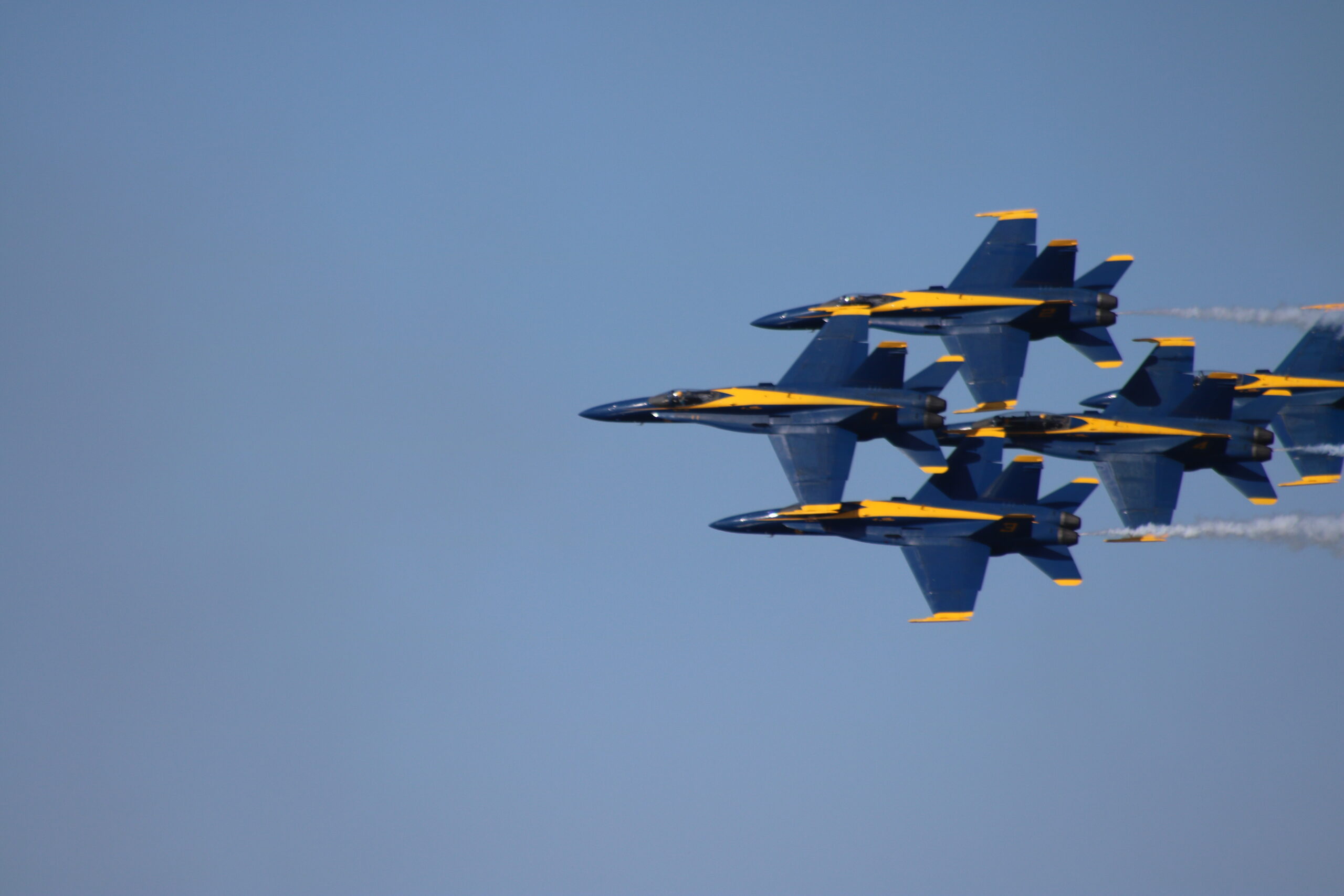 Four Blue Angels F/A-18 Super Hornets in diamond formation banked toward the camera against a clear blue sky, a fifth jet entering from the right edge