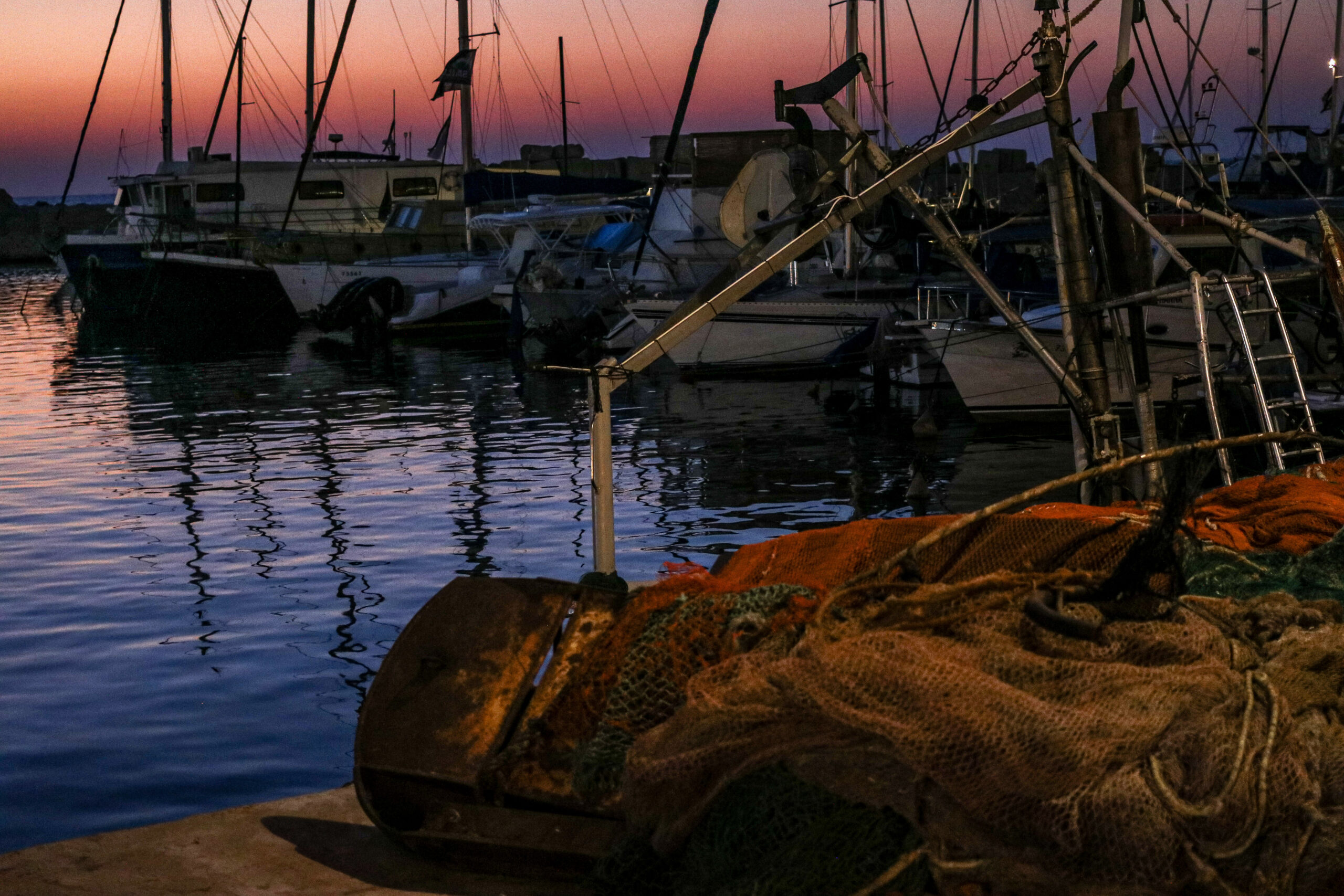 Fishing nets and a rusted drum on a harbor quay, with moored boats and masts against a pink and orange twilight sky