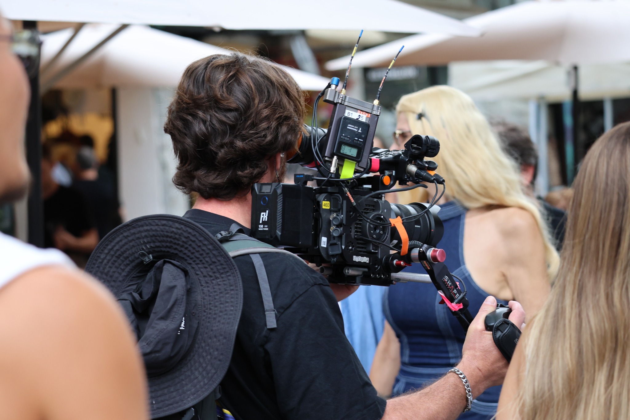 Camera operator filming in a crowd with a shoulder-mounted camera rig, external battery, wireless audio equipment and handgrip.