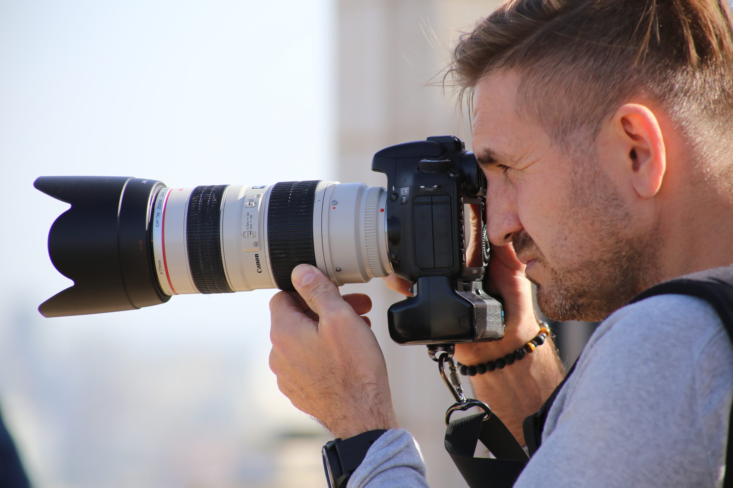 Photographer with his eye to a gripped camera body fitted with a white telephoto zoom against a bright sky