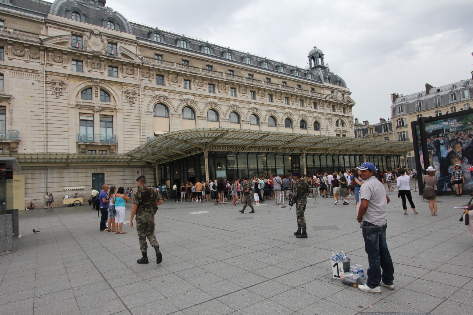 Armed soldiers patrol outside the Musée d’Orsay in Paris while tourists queue beneath the entrance canopy and a vendor stands beside bottled water.