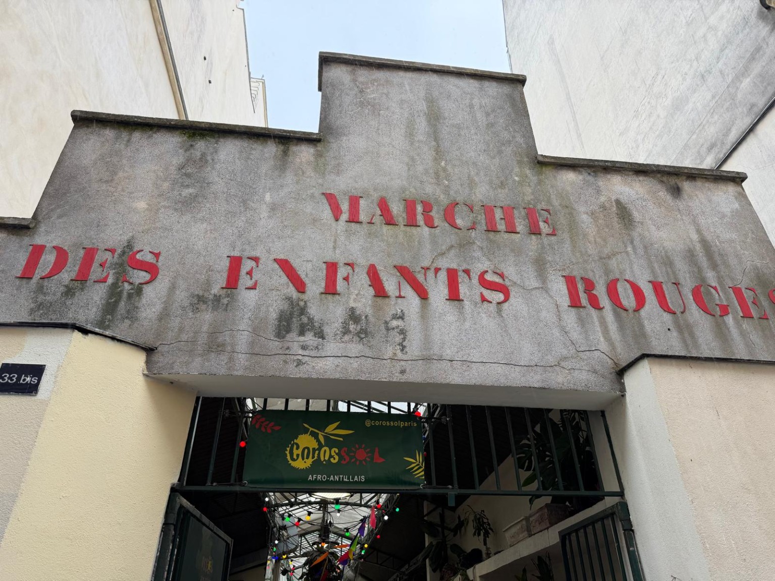 Weathered gray facade of the historic Marché des Enfants Rouges in Paris, with large red lettering, green iron entrance gates and a Corossol Afro-Caribbean food sign beneath the covered market.