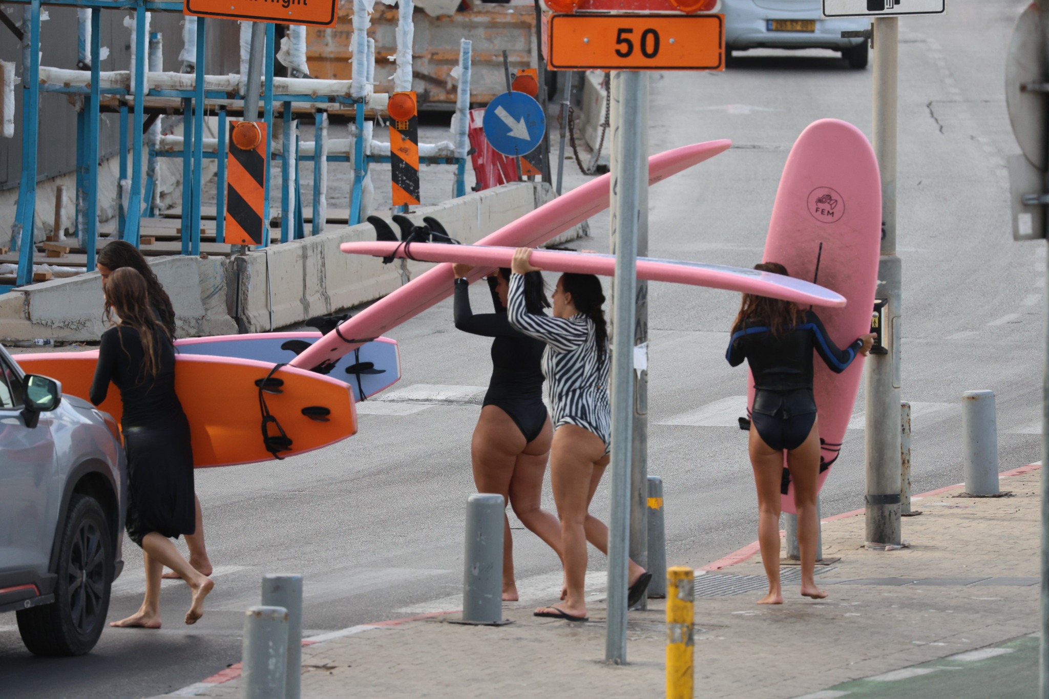 Surfers carrying pink and orange boards through a street lined with construction barriers, traffic signs and grey bollards